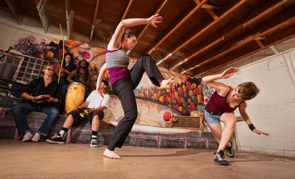 Two female capoeira performers sparring indoors while music plays