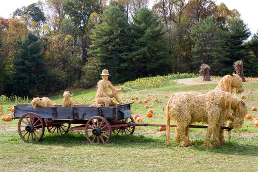Driver and animals made of straw sit in a old farm wagon pulled by straw horses in a pumpkin patch.
