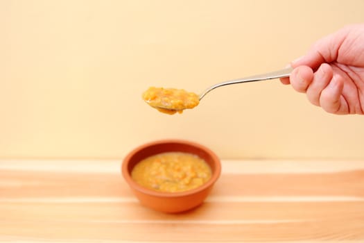 Hand holding spoonful of soup above a bowl on a wooden table with copy space