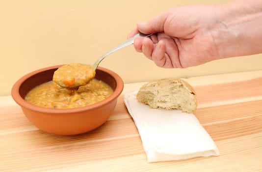 Hand lifts spoonful of vegetable soup from bowl with bread roll and napkin on table