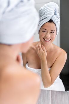portrait of a beautiful mature lady with towel looking at her skin in the mirror