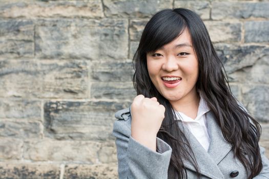 Portrait of successful young woman in front of a stone wall holding up fist