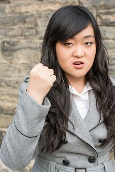 Portrait of young aggressive woman in front of a stone wall looking angry and holding fist