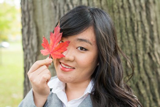 Autumn portrait of cute young woman in front of a maple tree holding a maple leaf and covering her eye