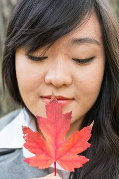 Autumn portrait of cute young woman in front of a maple tree holding a maple leaf with eyes closed