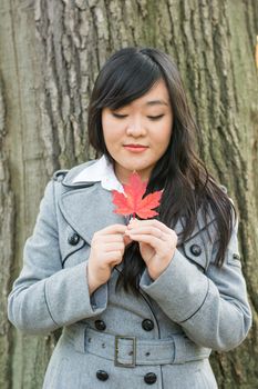 Autumn portrait of cute young woman in front of a maple tree holding a maple leaf with eyes closed