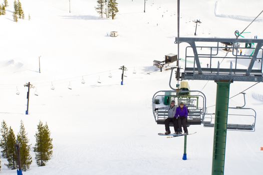 A couple of people ride the ski chair lift up the mountain together while sitting closely to each other having a fun time during a day of snowboarding in oregon.