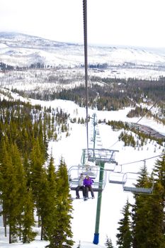 A couple of people ride the ski chair lift up the mountain together while sitting closely to each other having a fun time during a day of snowboarding in oregon.