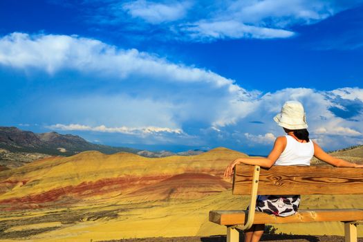Young woman sitting on the hill-top bench relaxing while watching the colorful Painted hills at John Day Fossil Beds National Monument, Oregon