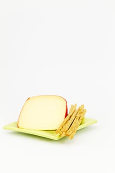 Slice of yellow cheese with red rind placed with breadsticks on green, square plate against white background; vertical image with large areas of copy space.