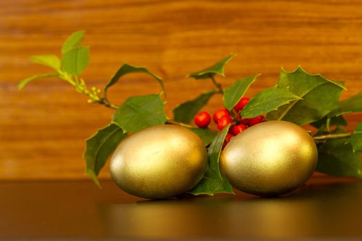 Two gold nest eggs with festive green holly sprigs and red berries placed on wood grain background;