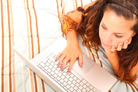 A young girl laying on the bed and surfing on the Internet with a Laptop.  