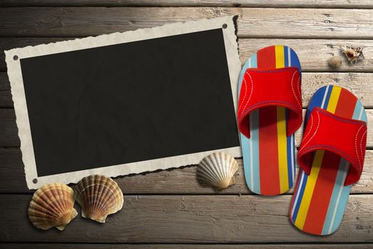 Aged photo frame with seashells on beach, colored sandals on wooden floor with sand