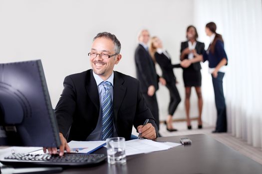 Confident happy businessman writing notes while using computer at office with colleagues in the background