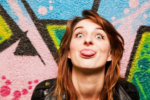 Stylish girl poking out her tongue to the camera against colorful graffiti wall.