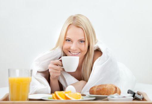 Beautiful woman drinking coffee in bed while eating breakfast