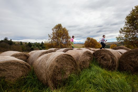 Young boy and girl play and run on cattle feed grass bales on mountain farm