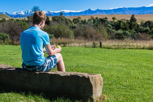 Boy teenager sitting on rock in morning light overlooking farm vegetation and distant mountains.