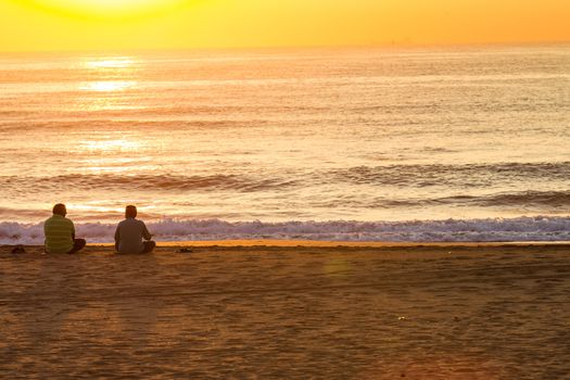 Two asian men sitting on beach sand watching sunrise over the ocean.