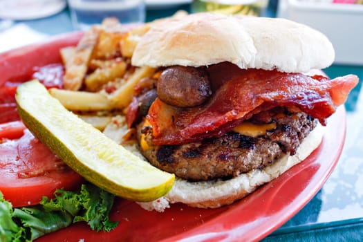 Gourmet burger with cheese mushrooms and thick cut bacon and a side of a pickle and some parmesan french fries.