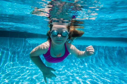 Young girl wearing goggles underwater in swimming pool.