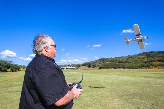 Mature man flies his remote control plane nearby over the field on a clear blue day