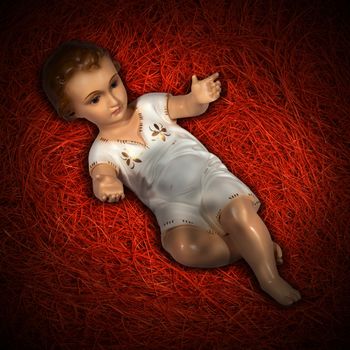 Baby Jesus figurine lying in manger on red straw background with shadows