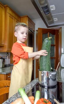 Cute child chef with apron cooking big zucchini and other vegetables in a pot on the kitchen