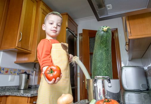 Cute child chef with apron cooking big zucchini and other vegetables in a pot on the kitchen