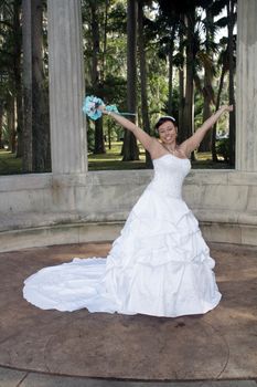 A lovely young bride outdoors wearing her wedding gown and holding her boquet, celebrates with her arms raised.