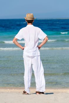 Back view a man with his hands on his hips in a white suit and hat standing on a tropical beach
