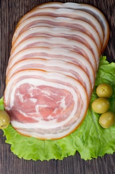 Slices of Delicious Smoked Ham Roll with Lettuce and Green Olives closeup on Wooden background