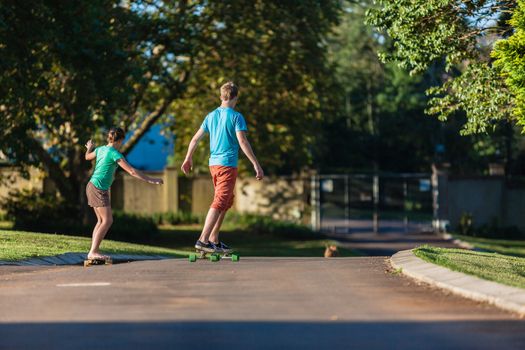 Girl and boy teenagers having fun skateboarding down small home tarred road in afternoon sunlight.