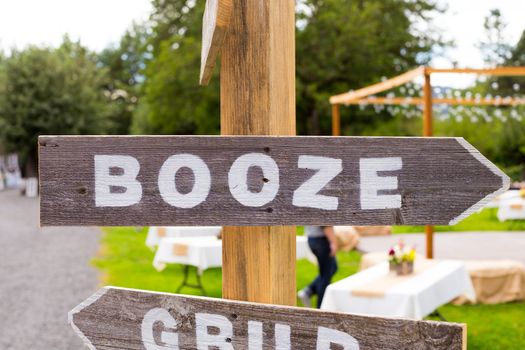 A handmade sign reads booze at an outdoor summer wedding pointing guests to the open bar.