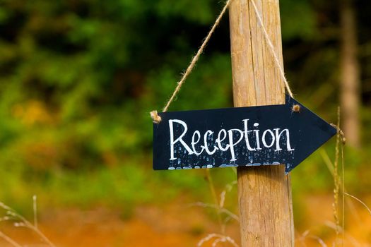A wedding reception sign is made from a small chalkboard and has been outside in the rain. The sign is an arrow hung with twine.