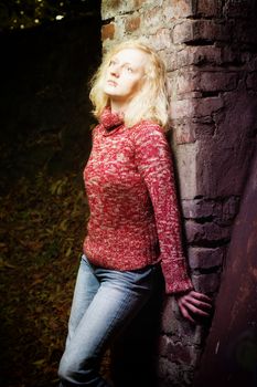 Beautiful young blond woman daydreaming at night standing outdoors with her back against a brick wall staring up into the darkness, she is illuminated