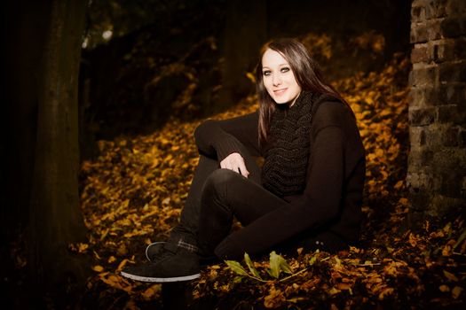 Beautiful woman in autumn leaves sitting outside in the darkness on the ground alongside a brick wall, with copyspace