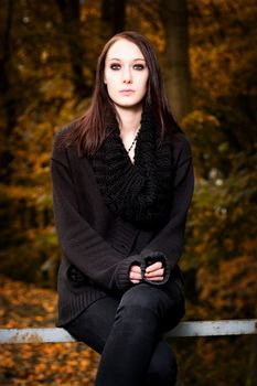 Vertical portrait of a mysterious beautiful young brunette woman sitting on a bench in the forest, wearing black clothes, in Autumn