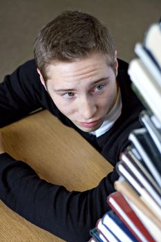 A frustrated student looks up at the high pile of textbooks he has to go through to do his homework assignment. Shallow depth of field.
