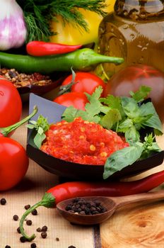 Arrangement of Bruschetta Sauce in Black Bowl with Ingredients, Spices and Black Peppercorn in Wooden Spoon closeup on Wooden background