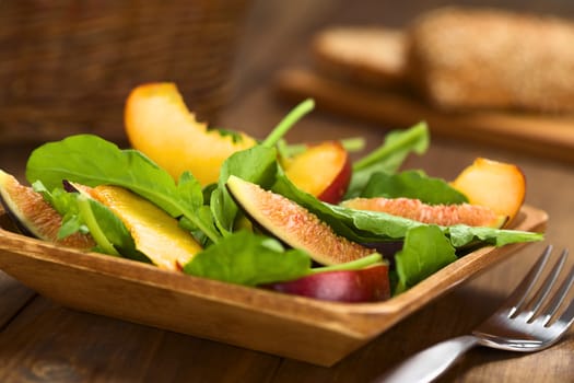 Fresh vegetarian salad made of fig, nectarine, spinach, cucumber and lettuce on wooden plate with wholewheat bread in the back (Selective Focus, Focus on the fig slice in the front)   