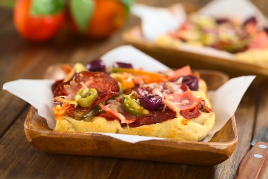 Fresh homemade pizza piece with tomato sauce, ham, salami, olives, bell pepper and cheese on top, served on sandwich paper on wooden plate (Selective Focus, Focus on the front of the olive slices)