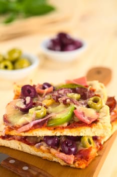 Fresh homemade pizza pieces with tomato sauce, ham, salami, olives, bell pepper and cheese on top on wooden board with knife (Selective Focus, Focus one third into the image)
