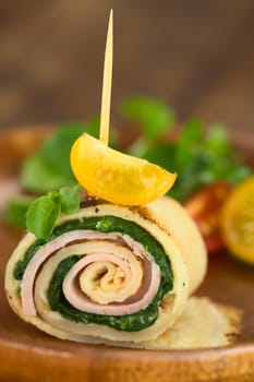 Crepe roll as finger food filled with spinach and ham garnished with cherry tomato and watercress served on wooden plate (Selective Focus, Focus on the upper part of the crepe roll and on the front of the cherry tomato on top) 