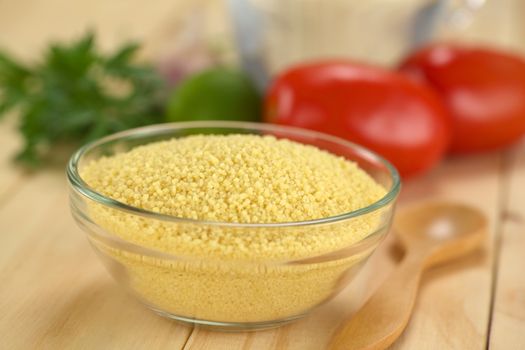 Raw couscous in glass bowl with ingredients in the back (Selective Focus, Focus one third into the couscous)