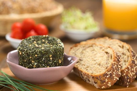 Goat cheese covered with herbs in bowl, chives and slices of wholegrain bread on wooden board (Selective Focus, Focus on the front of the cheese)