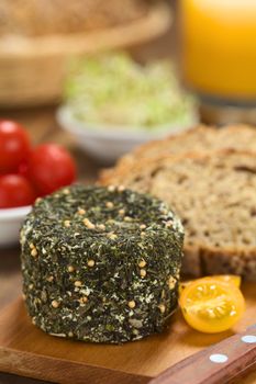 Goat cheese covered with herbs, yellow cherry tomato and slices of wholegrain bread on wooden board (Selective Focus, Focus on the upper edge of the cheese)