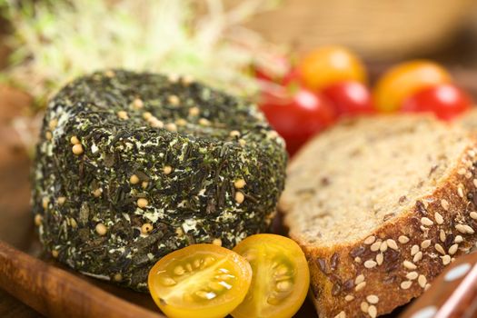 Goat cheese covered with herbs, yellow cherry tomato and slices of wholegrain bread on wooden plate (Selective Focus, Focus on the upper edge of the cheese)