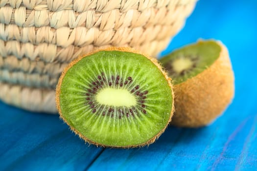 Half kiwi fruits on blue wood with woven basket behind (Selective Focus, Focus on the half kiwi)