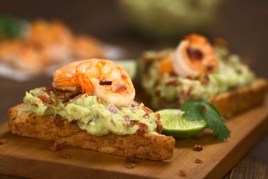 Wholegrain toast bread slices with guacamole, fried shrimp and fried bacon pieces on wooden board (Selective Focus, Focus on the front of the shrimp on the first bread) 